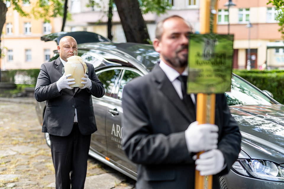 Pracownicy firmy Archon w strojach galowych uczestniczą w ceremonii pogrzebowej przed kaplicą. Jeden z nich niesie urnę, a drugi tabliczkę, stoją przy karawanie na tle zieleni i budynków.