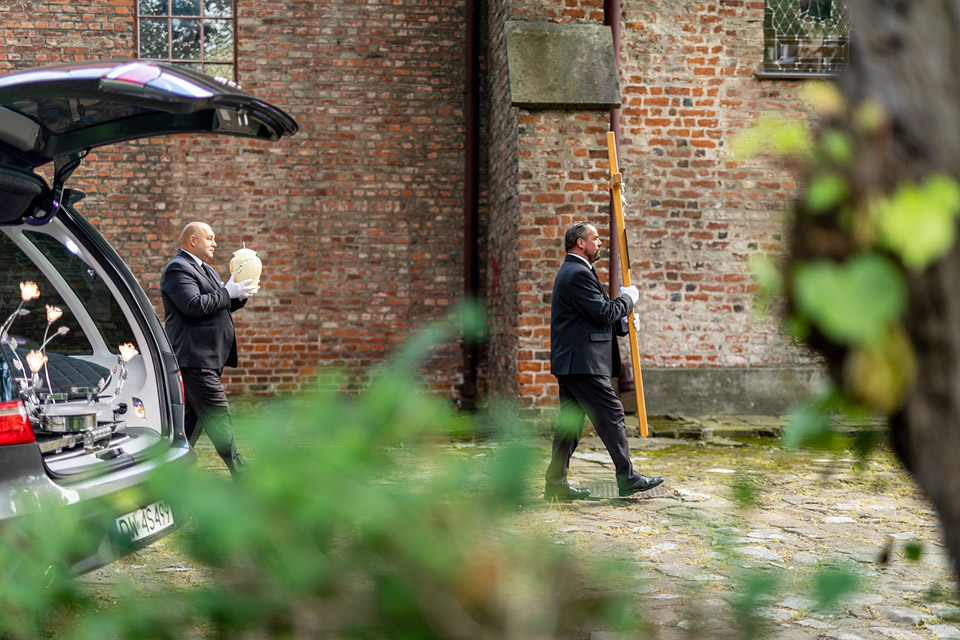 Dwaj pracownicy ubrani w garnitury niosą urnę oraz krzyż w pobliżu kaplicy. Tło to ceglana ściana. Widoczny jest otwarty samochód.