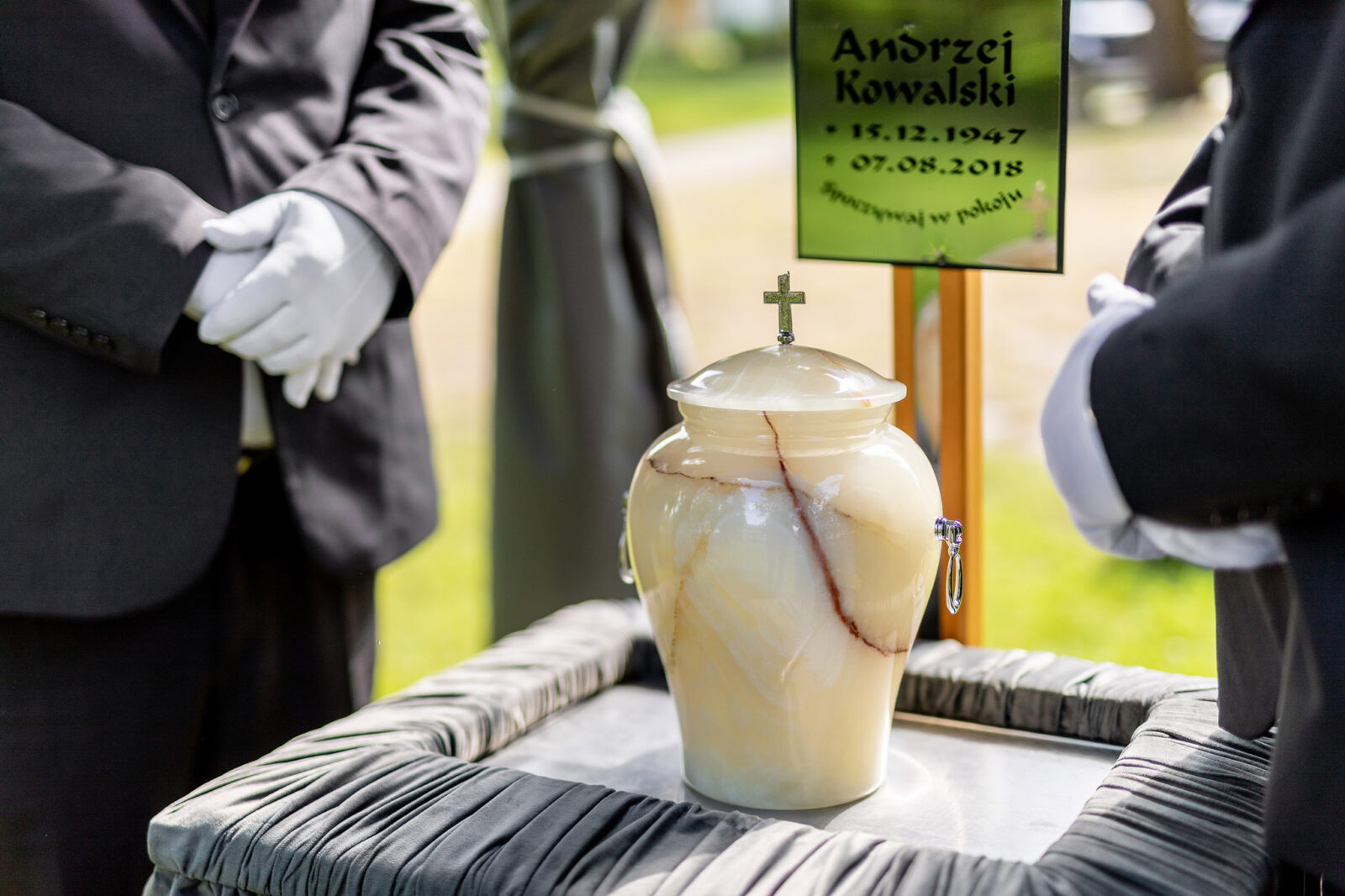 Zbliżenie na urnę podczas ceremonii pogrzebowej organizowanej przez firmę Archon. Dwie osoby w rękawiczkach stoją obok urny na pokrytym materiałem stoliku, w tle tablica z imieniem Andrzej Kowalski i datami jego życia.