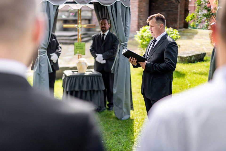 Pracownicy firmy Archon podczas ceremonii pogrzebowej w kaplicy, mężczyzna czyta z notesu obok urny i krzyża.