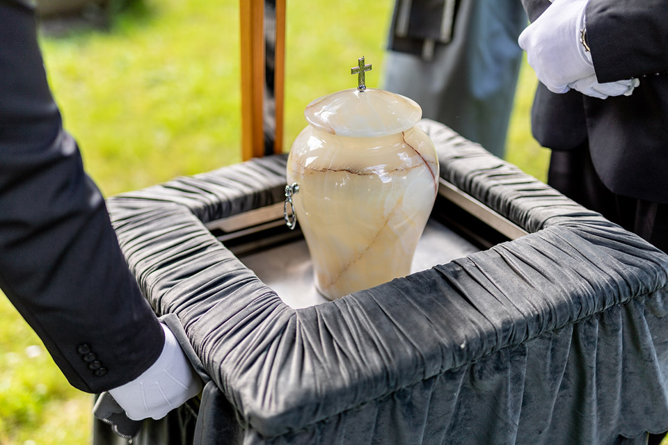 Pracownicy firmy Archon podczas ceremonii pogrzebowej, trzymający urnę ozdobioną krzyżem na aksamitnym podkładzie.