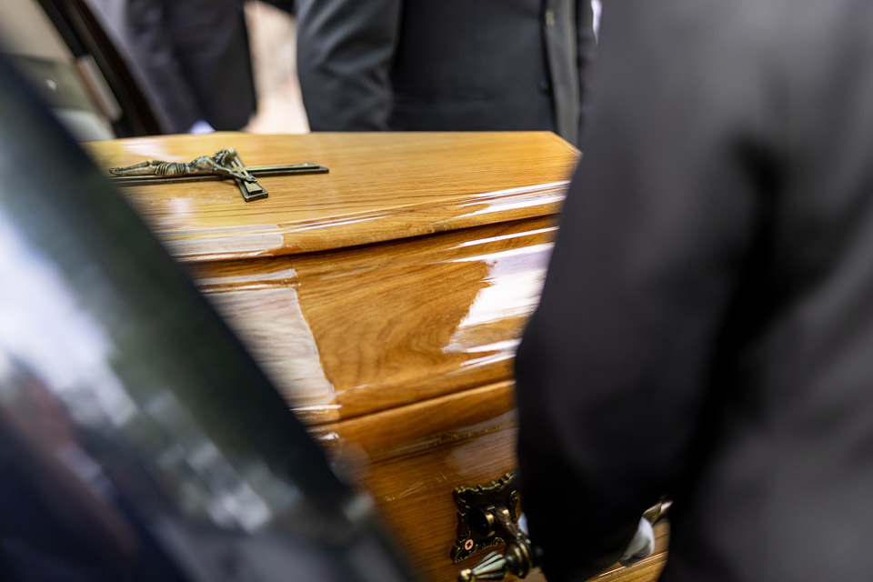 Pracownicy firmy Archon niosą trumnę do kaplicy. Zbliżenie na drewnianą trumnę z krzyżem.