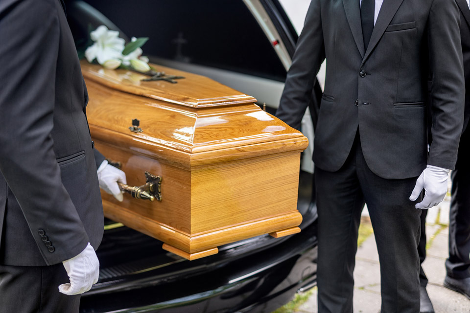 Pracownicy firmy pogrzebowej Archon transportują trumnę do karawanu.