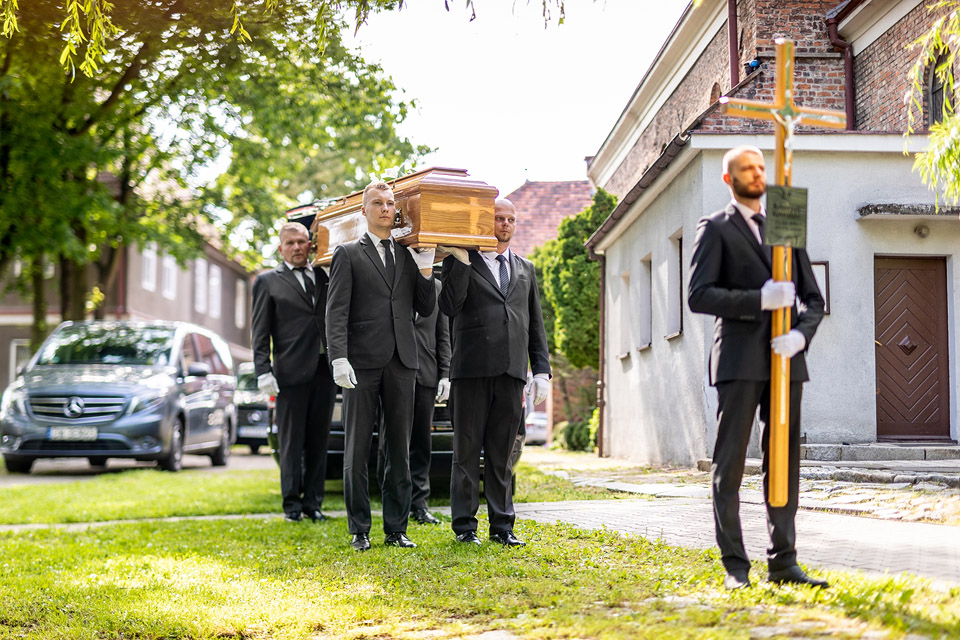 Pracownicy zakładu pogrzebowego Archon w eleganckich garniturach niosą trumnę podczas procesji przed kaplicą, z przodu osoba z krzyżem.