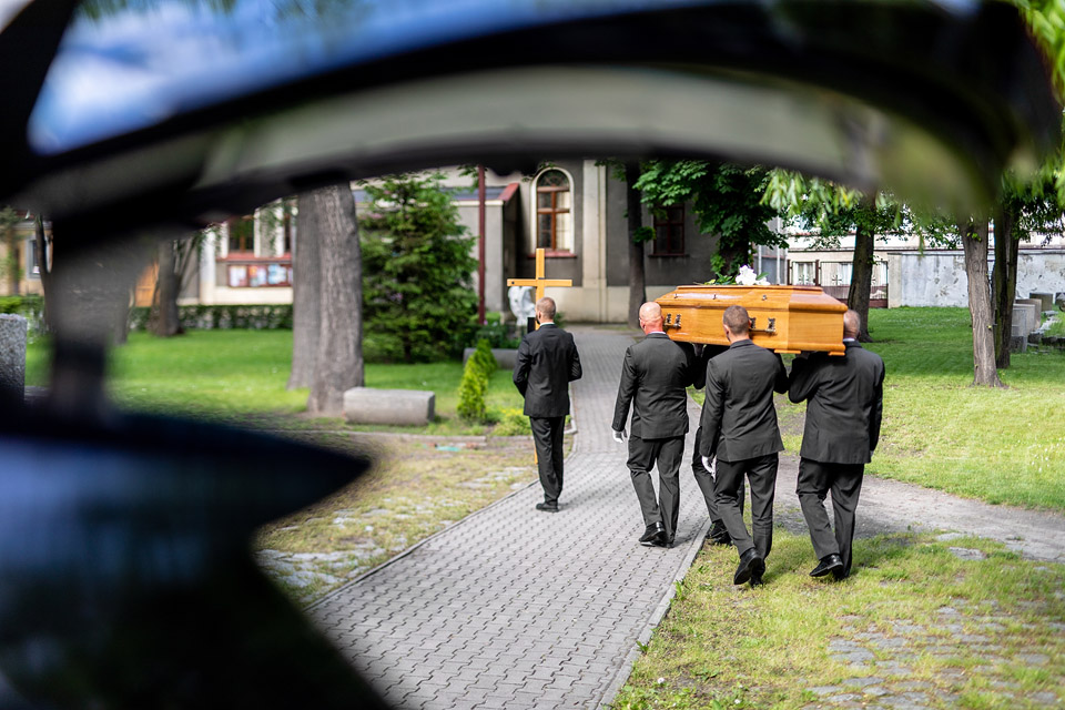 Czterej mężczyźni ubrani w garnitury niosą trumnę w kierunku kaplicy, otoczeni zielenią.