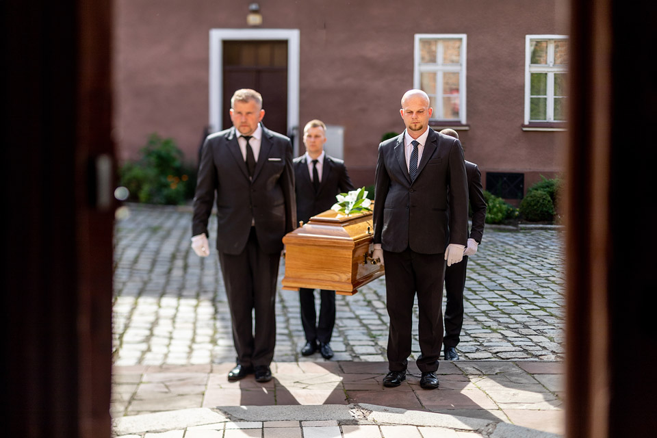 Zespół pracowników firmy Archon niesie trumnę na dziedziniec przed kaplicą. Czterech mężczyzn ubranych w eleganckie garnitury prowadzi ceremonialny pochód.